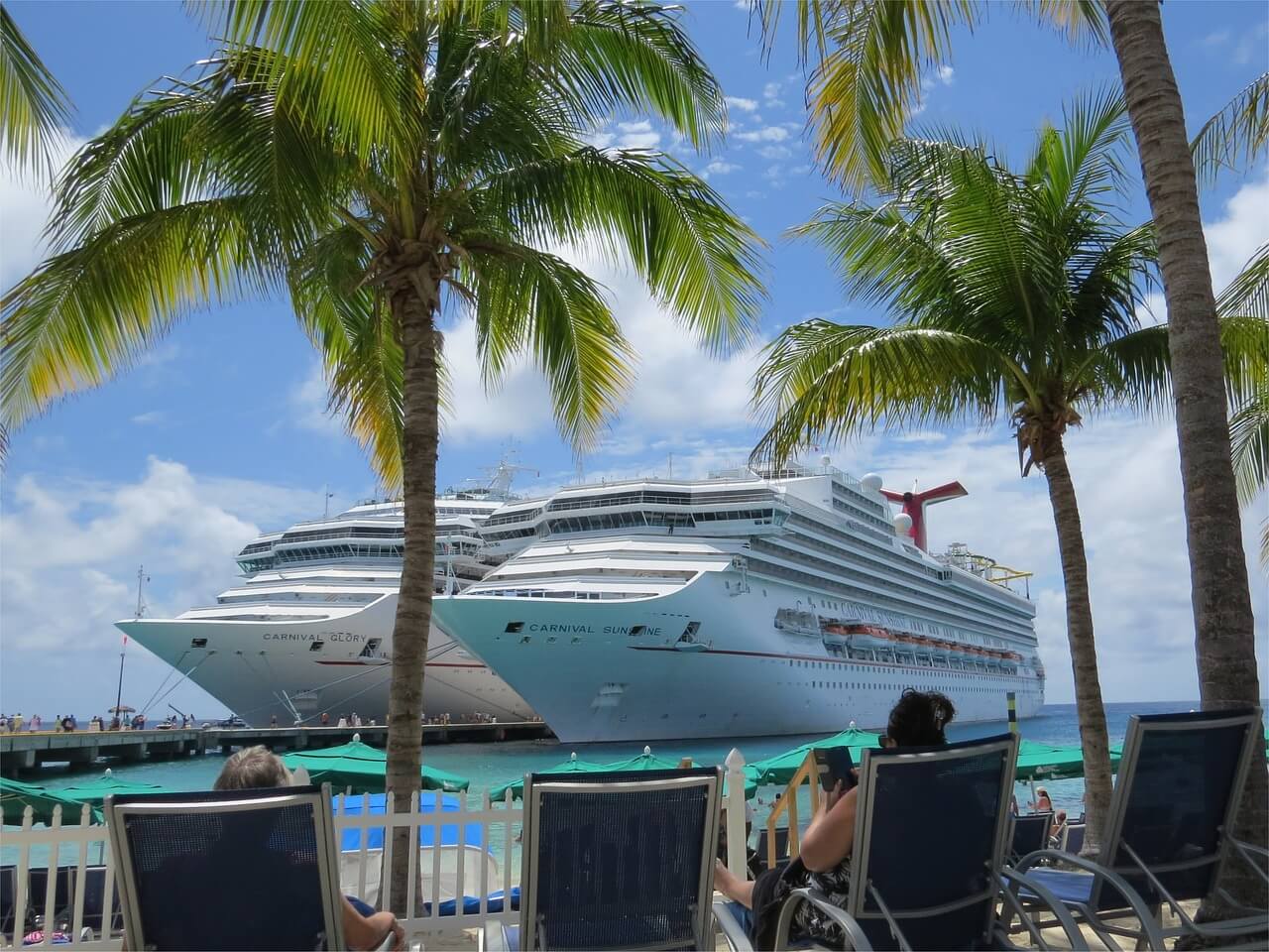 A cruise ship seen past palm trees from a beach bar lined with deck chairs