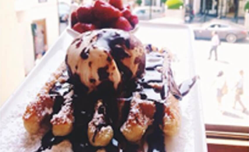 A Belgian waffle heaped with chocolate sauce and cream on a plate by a window