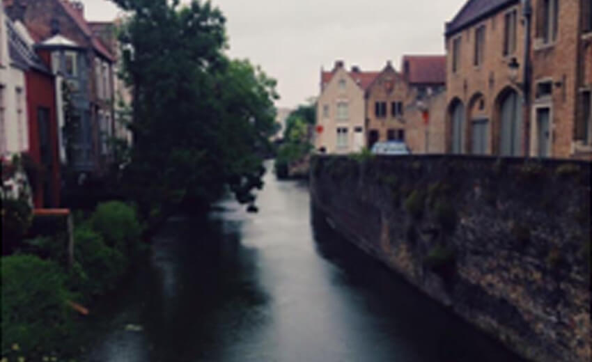 A canal in Bruges running between old houses and a stone wall, trees overhanging the water