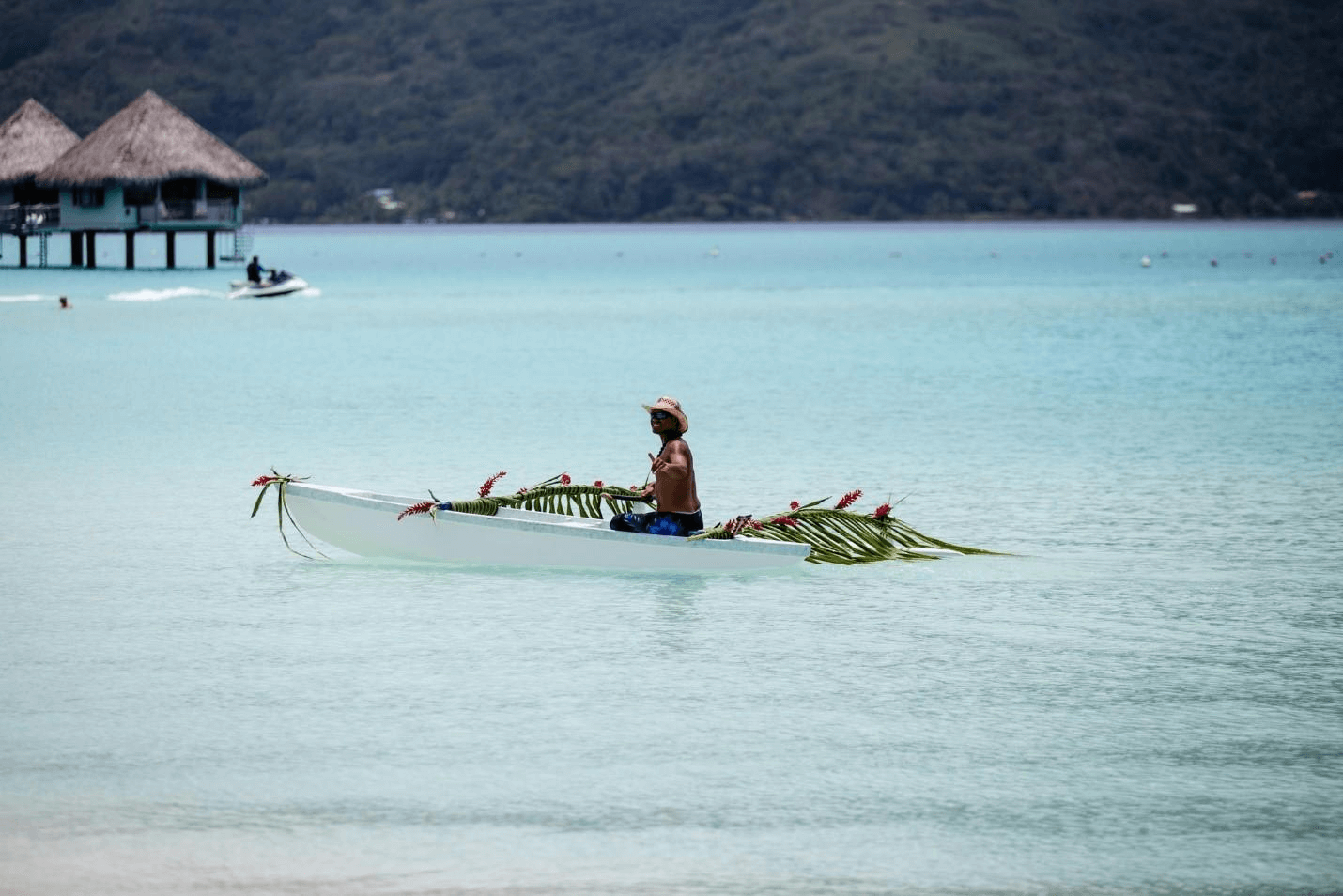 A man paddling an outrigger canoe loaded with palm fronds across a pale lagoon