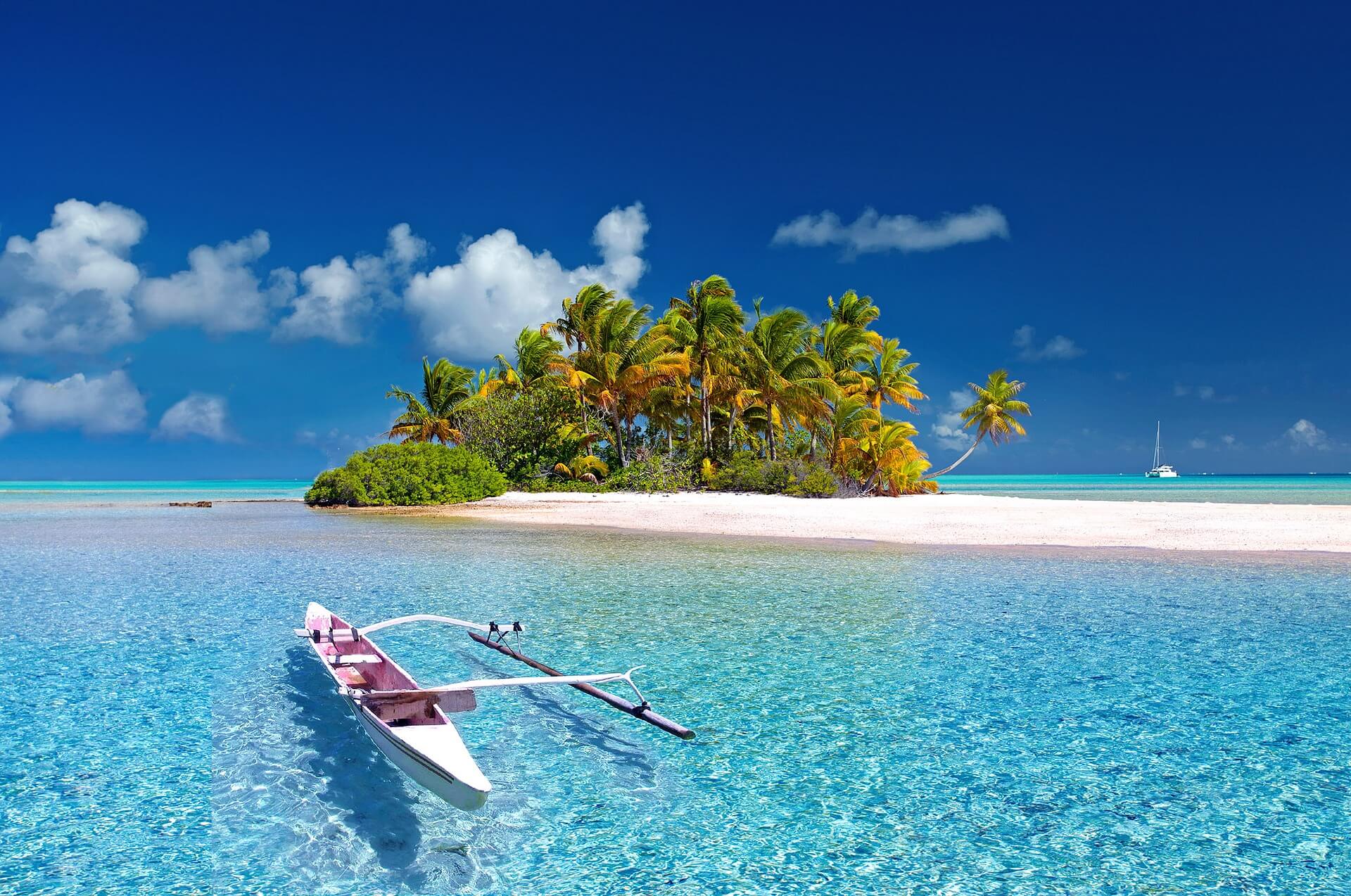 A small palm-covered island on a white sandbar, an outrigger canoe in clear shallow water