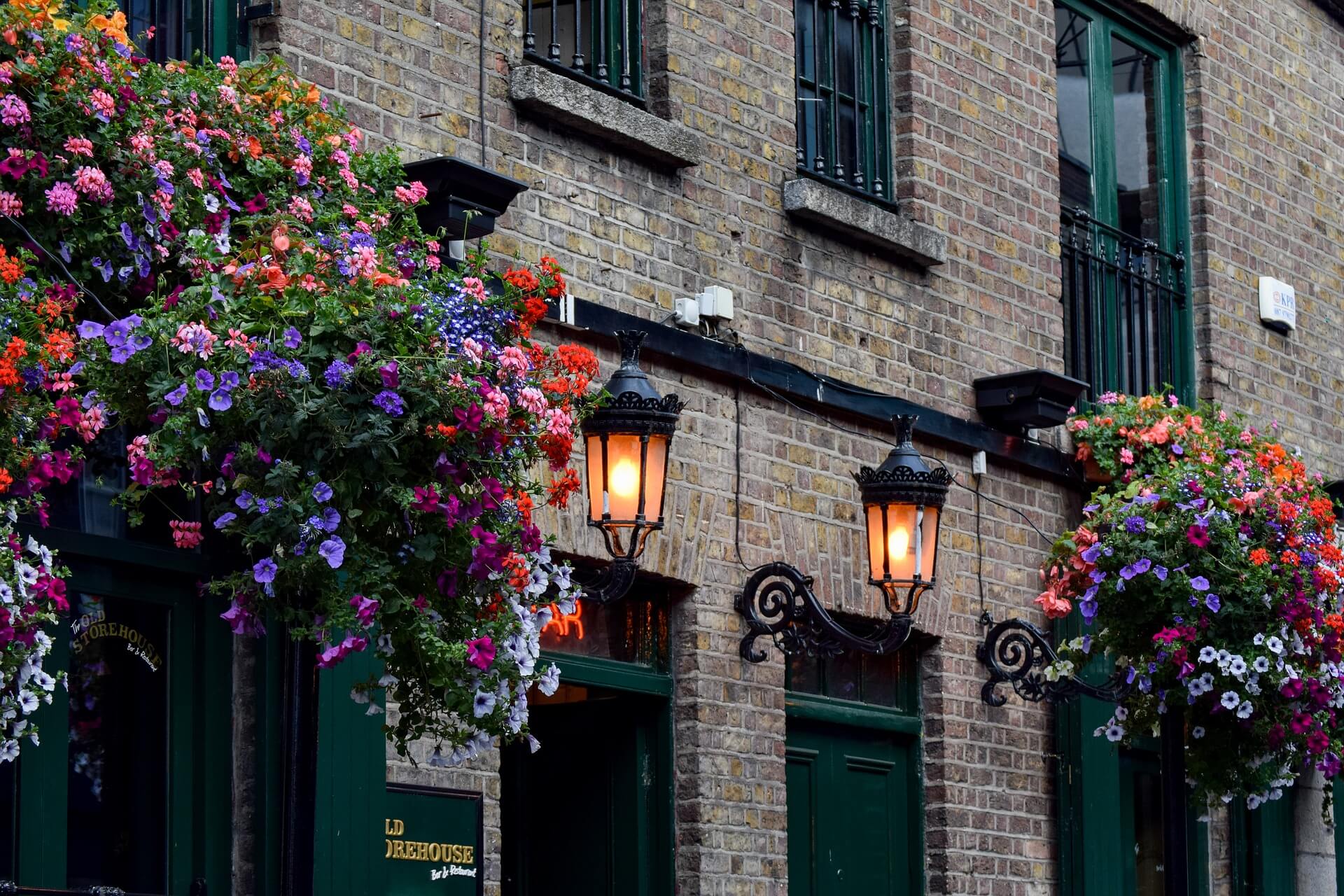The front of an Irish pub in brick and green paintwork, two lit lanterns and hanging flower baskets