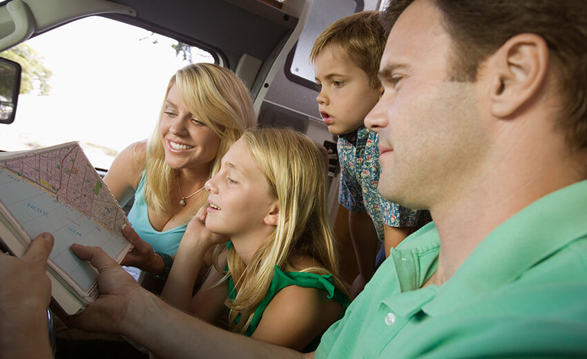 A family of four in a car, two adults in front and two children behind, looking at a map